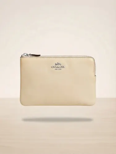 cartera de mano (wristlet) de la marca Coach, modelo 'Corner Zip', en un elegante color beige claro o crema.